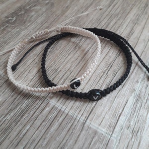 Peut inclure: Deux bracelets d'amitié, un noir et un beige, avec un charme en forme de lettre "V" noire sur chacun.