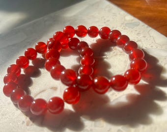 May include: Three red beaded bracelets. Each bracelet is made of round, polished beads. The bracelets are arranged on a light-colored, textured surface. The beads have a translucent quality, and the color is a deep, rich red.