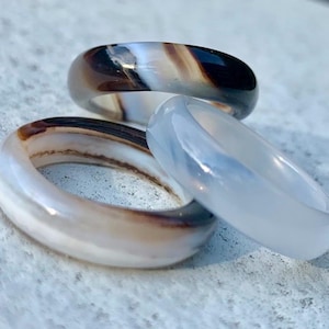 Puede incluir: Tres anillos de piedras preciosas en tonos marrón, blanco y azul claro. Los anillos son lisos y pulidos, con forma redondeada. Los anillos se exhiben sobre una superficie texturizada de color claro.