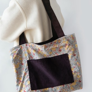 May include: A floral print tote bag with a dark purple corduroy pocket and straps. The bag features a pattern of yellow and red flowers on a light blue background. The straps and pocket are a deep purple color.