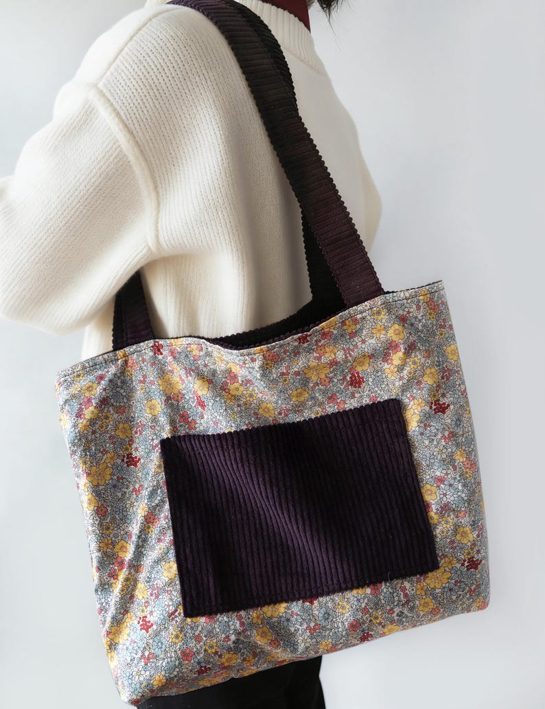May include: A floral print tote bag with a dark purple corduroy pocket and straps. The bag features a pattern of yellow and red flowers on a light blue background. The straps and pocket are a deep purple color.