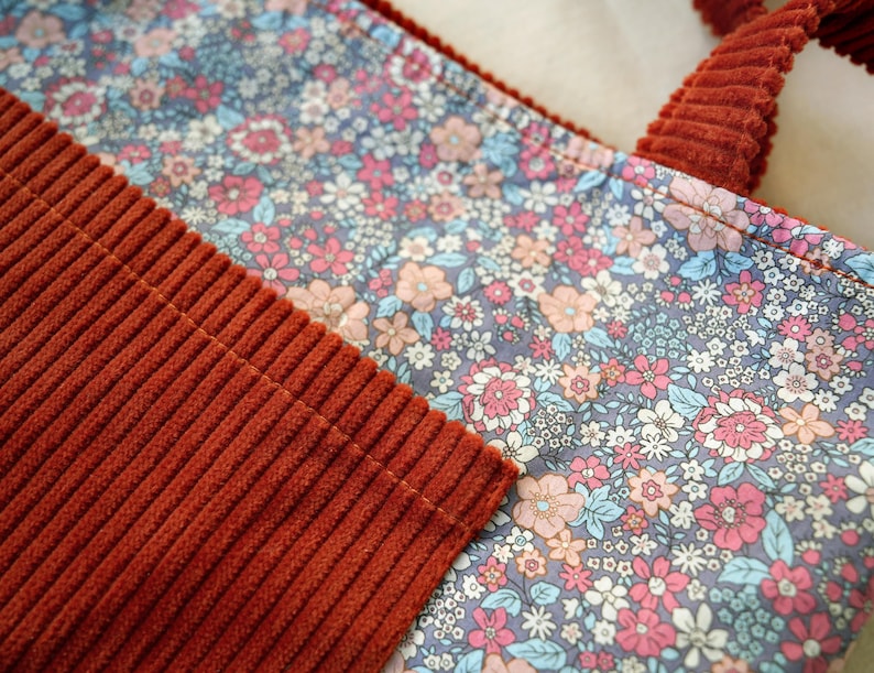 May include: Close-up of a floral patterned fabric tote bag with rust-colored corduroy handles and accents. The floral pattern features pink, blue, and peach flowers on a gray background. The corduroy has a ribbed texture.
