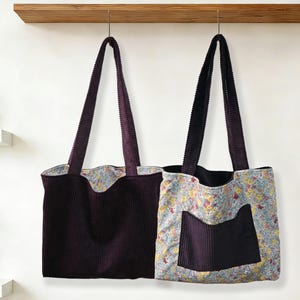 May include: Two corduroy tote bags hanging against a white wall. One is solid dark purple, the other has a floral print with a dark purple pocket. Both have matching dark purple corduroy straps.