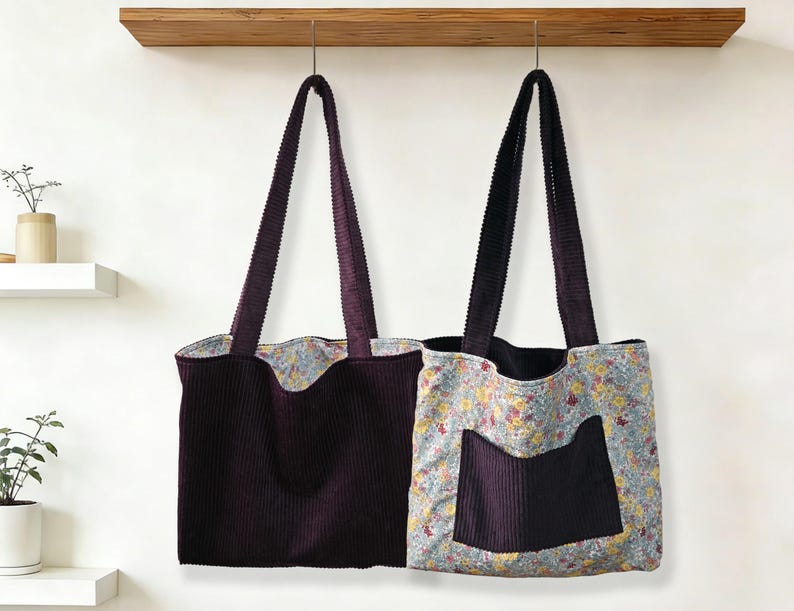 May include: Two corduroy tote bags hanging against a white wall. One is solid dark purple, the other has a floral print with a dark purple pocket. Both have matching dark purple corduroy straps.
