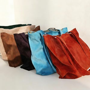 May include: A collection of six corduroy tote bags in various colors, including green, tan, brown, purple, blue, and orange. The bags are arranged in a row, showcasing their textured fabric and simple design. Each bag has a shoulder strap.