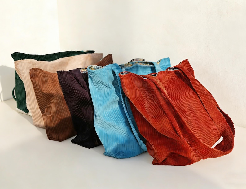 May include: A collection of six corduroy tote bags in various colors, including green, tan, brown, purple, blue, and orange. The bags are arranged in a row, showcasing their textured fabric and simple design. Each bag has a shoulder strap.
