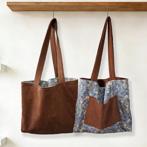 May include: Two corduroy tote bags hanging against a white wall. One is solid brown, the other has a floral pattern with a brown pocket. Both have long brown straps and are hanging from a wooden shelf.