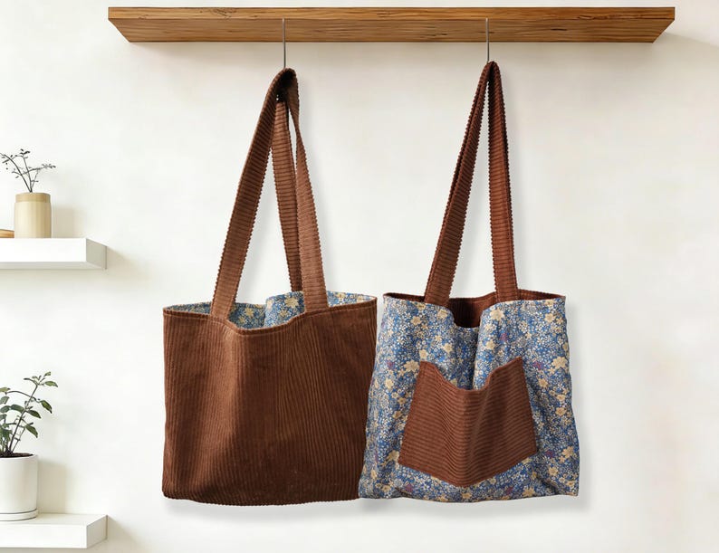 May include: Two corduroy tote bags hanging against a white wall. One is solid brown, the other has a floral pattern with a brown pocket. Both have long brown straps and are hanging from a wooden shelf.
