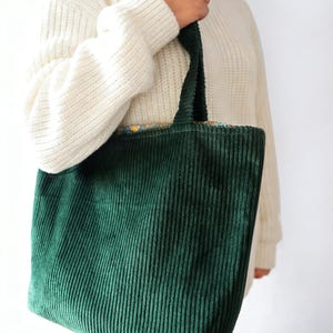 May include: A green corduroy tote bag with matching handles. The bag is held by a person wearing a cream-colored sweater. The tote bag has a rectangular shape and a textured surface.