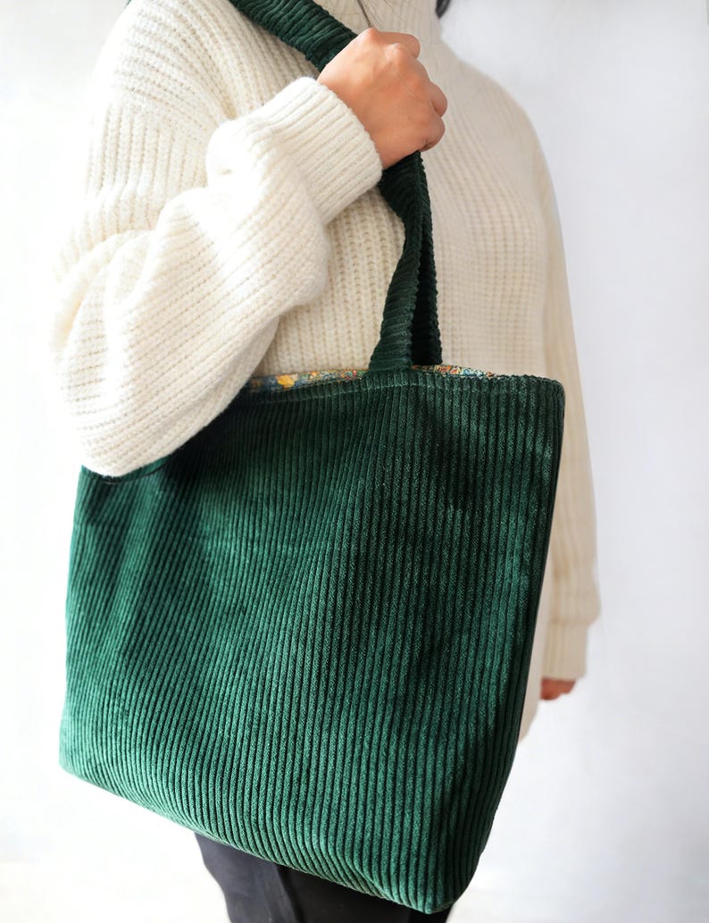 May include: A green corduroy tote bag with matching handles. The bag is held by a person wearing a cream-colored sweater. The tote bag has a rectangular shape and a textured surface.
