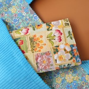Puede incluir: Una cartera con estampado floral con flores rojas, rosas y blancas sobre un fondo amarillo. La cartera est&aacute; rodeada de tela de pana azul y una tela floral con flores azules y amarillas.