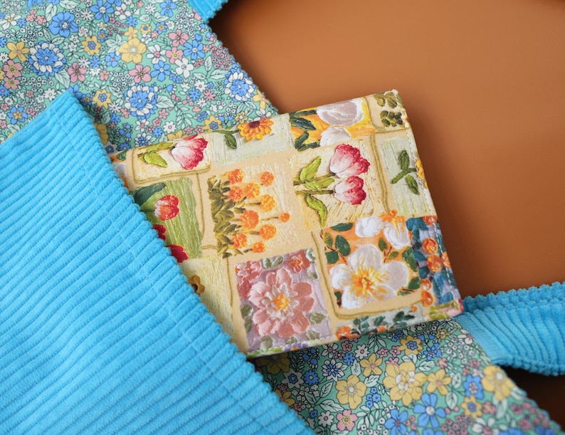 Puede incluir: Una cartera con estampado floral con flores rojas, rosas y blancas sobre un fondo amarillo. La cartera est&aacute; rodeada de tela de pana azul y una tela floral con flores azules y amarillas.