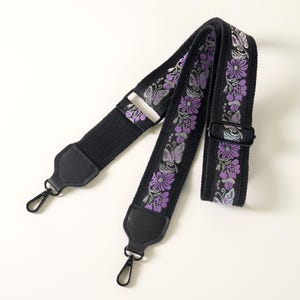 May include: A black adjustable bag strap featuring a woven floral and butterfly pattern in shades of purple and silver. The strap includes black leather-like end tabs, black metal swivel lobster clasps, and a black metal sliding buckle adjuster.
