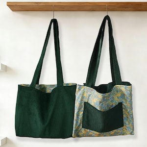 May include: Two green corduroy tote bags hanging from a wooden shelf. One bag is solid green, while the other has a floral patterned lining and a green corduroy pocket. Both have long, green straps.