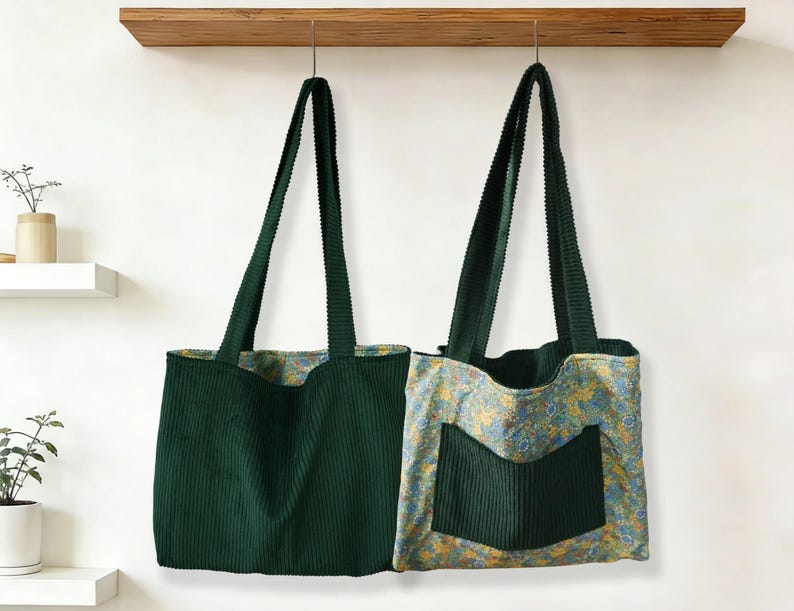 May include: Two green corduroy tote bags hanging from a wooden shelf. One bag is solid green, while the other has a floral patterned lining and a green corduroy pocket. Both have long, green straps.