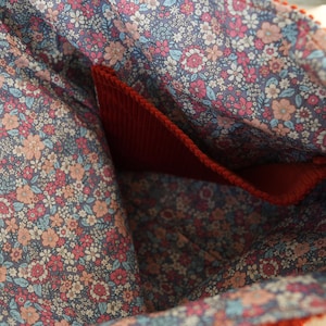May include: Close-up of a red corduroy bag interior with a floral patterned lining. The lining features a blue background with pink, peach, and white flowers. A red pocket is visible inside the bag.
