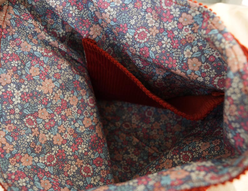May include: Close-up of a red corduroy bag interior with a floral patterned lining. The lining features a blue background with pink, peach, and white flowers. A red pocket is visible inside the bag.