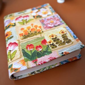 Può includere: Un libro con copertina a fantasia floreale. La copertina presenta un design patchwork con vari fiori colorati, tra cui tulipani, margherite e rose, su sfondo beige. I bordi del libro sono visibili, mostrando le pagine bianche.