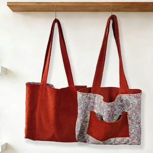 May include: Two corduroy tote bags hanging against a white wall. One is solid rust-colored, the other has a floral print lining and pocket. Both have long, matching rust-colored straps.