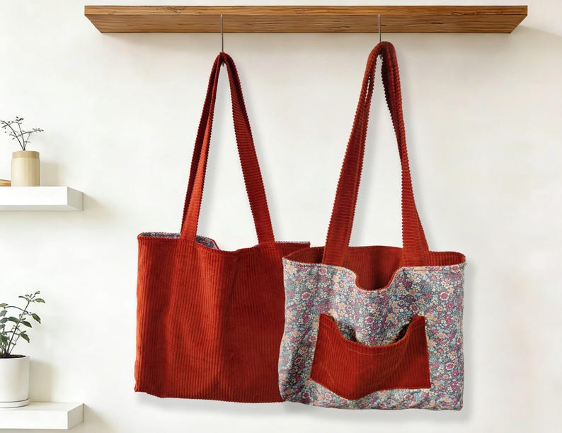 May include: Two corduroy tote bags hanging against a white wall. One is solid rust-colored, the other has a floral print lining and pocket. Both have long, matching rust-colored straps.