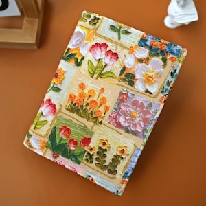 Puede incluir: Un diario con estampado floral con un fondo beige y cuadrados de flores coloridas. La cubierta presenta flores rojas, naranjas y blancas con hojas verdes. El diario es rectangular y parece estar cerrado.