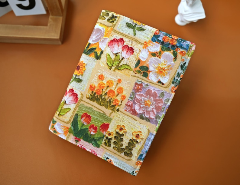 Puede incluir: Un diario con estampado floral con un fondo beige y cuadrados de flores coloridas. La cubierta presenta flores rojas, naranjas y blancas con hojas verdes. El diario es rectangular y parece estar cerrado.