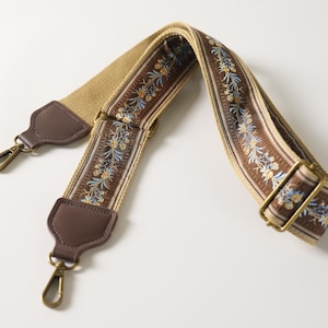 Könnte beinhalten: Ein beigefarbener und brauner, verstellbarer Taschenriemen mit einem Blumenmuster aus blauen und gelben Blüten. Der Riemen verfügt über dunkelbraune Kunstleder-Endstücke, schwenkbare Karabinerhaken aus messingfarbenem Metall und eine passende Metallschnalle.
