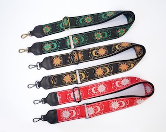 Op de afbeelding: Drie verstelbare tasriemen met draaibare haken en zwarte lederen uiteinden. De bovenste riem is zwart met groene en gele zon- en maanmotieven. De middelste riem is zwart met gouden ontwerpen. De onderste riem is rood met witte zon- en maanpatronen.