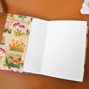 Puede incluir: Cuaderno abierto con una cubierta de dise&ntilde;o floral con tulipanes rojos y rosas, flores amarillas y hojas verdes. Las p&aacute;ginas del cuaderno est&aacute;n en blanco y son blancas. La cubierta tiene un fondo color crema.