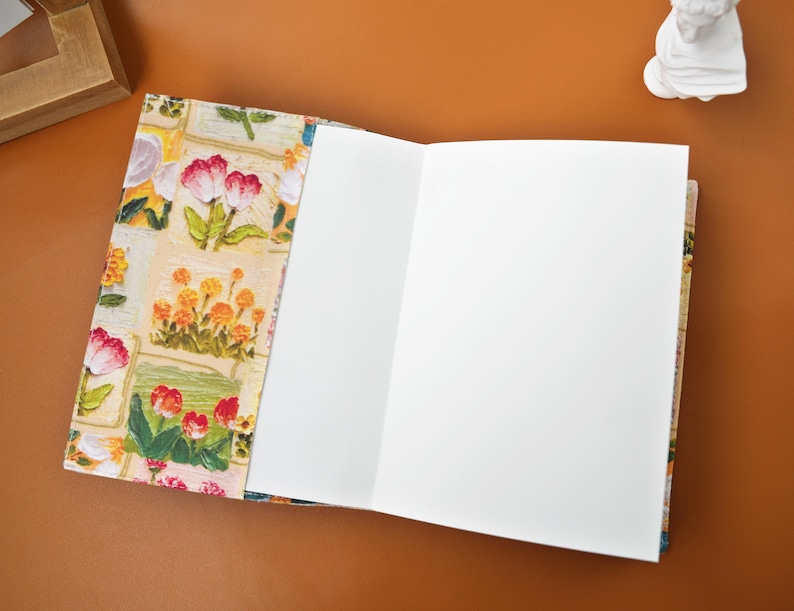 Puede incluir: Cuaderno abierto con una cubierta de dise&ntilde;o floral con tulipanes rojos y rosas, flores amarillas y hojas verdes. Las p&aacute;ginas del cuaderno est&aacute;n en blanco y son blancas. La cubierta tiene un fondo color crema.