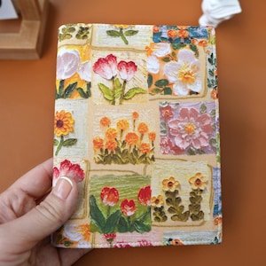 Puede incluir: Una cubierta de cuaderno con un dise&ntilde;o de patchwork floral. La cubierta muestra varias ilustraciones de flores en rojo, naranja, amarillo y blanco. La cubierta est&aacute; sujeta por una mano, destacando la obra de arte detallada y la superficie texturizada.