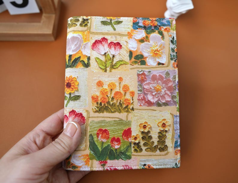 Puede incluir: Una cubierta de cuaderno con un dise&ntilde;o de patchwork floral. La cubierta muestra varias ilustraciones de flores en rojo, naranja, amarillo y blanco. La cubierta est&aacute; sujeta por una mano, destacando la obra de arte detallada y la superficie texturizada.