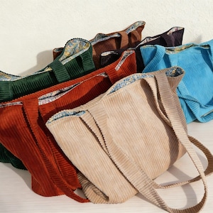May include: A collection of corduroy tote bags in various colors, including green, rust, tan, brown, and blue. Each bag features a matching corduroy strap and a patterned interior lining. The bags are arranged in a slightly overlapping display.
