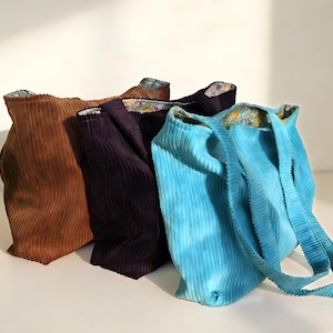 May include: Four corduroy tote bags in shades of brown, purple, and turquoise. The bags have a ribbed texture and floral patterned lining. The turquoise bag has long straps. The bags are standing upright.