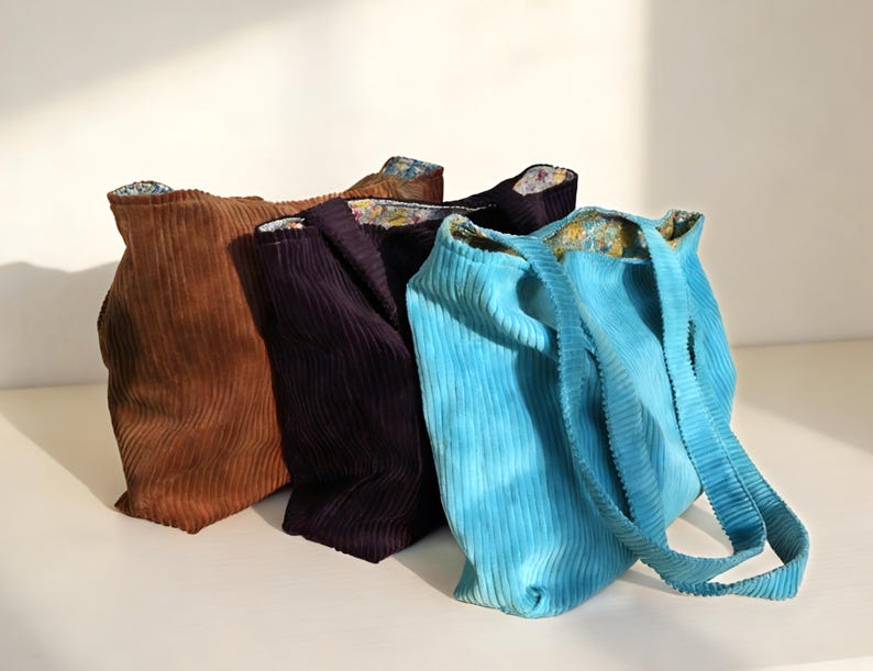 May include: Four corduroy tote bags in shades of brown, purple, and turquoise. The bags have a ribbed texture and floral patterned lining. The turquoise bag has long straps. The bags are standing upright.