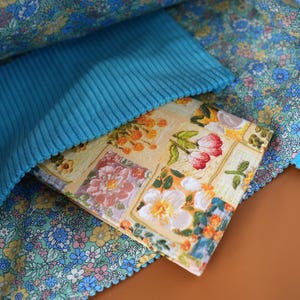 Puede incluir: Un bolso tote con estampado floral con un bolsillo de pana turquesa. El interior del bolso presenta una tela floral azul claro y un libro con estampado floral. El libro tiene un fondo amarillo con flores rosas, blancas y naranjas.