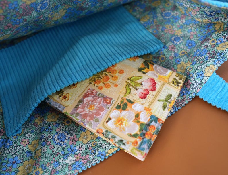 Puede incluir: Un bolso tote con estampado floral con un bolsillo de pana turquesa. El interior del bolso presenta una tela floral azul claro y un libro con estampado floral. El libro tiene un fondo amarillo con flores rosas, blancas y naranjas.