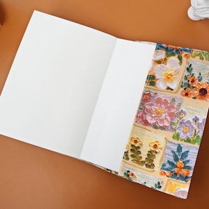 Puede incluir: Un cuaderno abierto con p&aacute;ginas blancas en blanco. La portada presenta un colorido dise&ntilde;o floral con cuadrados de flores en tonos de rosa, naranja, amarillo y morado. El cuaderno est&aacute; sobre una superficie marr&oacute;n.