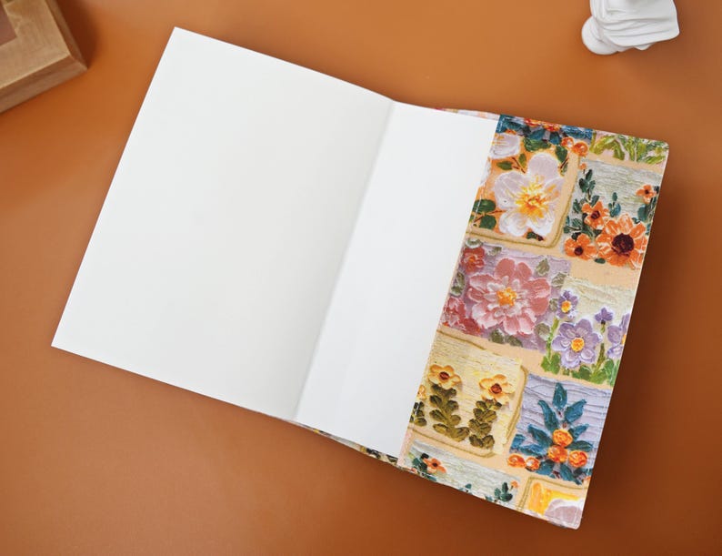 Puede incluir: Un cuaderno abierto con p&aacute;ginas blancas en blanco. La portada presenta un colorido dise&ntilde;o floral con cuadrados de flores en tonos de rosa, naranja, amarillo y morado. El cuaderno est&aacute; sobre una superficie marr&oacute;n.