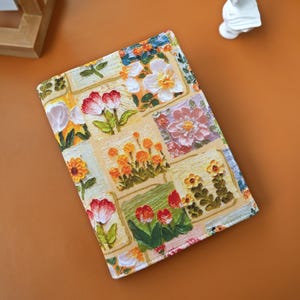 Puede incluir: Una cubierta de libro rectangular con un dise&ntilde;o floral de patchwork. El dise&ntilde;o presenta varias flores rojas, naranjas, amarillas y blancas sobre un fondo crema. El dise&ntilde;o de la tela presenta una mezcla de colores y estilos.