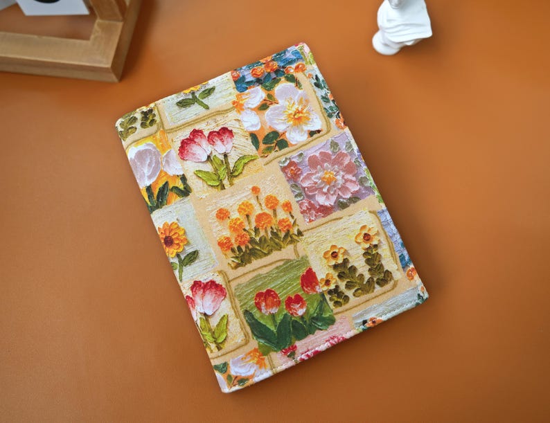 Puede incluir: Una cubierta de libro rectangular con un dise&ntilde;o floral de patchwork. El dise&ntilde;o presenta varias flores rojas, naranjas, amarillas y blancas sobre un fondo crema. El dise&ntilde;o de la tela presenta una mezcla de colores y estilos.