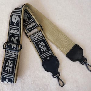 Könnte beinhalten: Ein verstellbarer Taschenriemen mit einer beigen, gewebten Stoffbasis und einem schwarz-weißen Muster. Der Riemen verfügt über schwarze Metallbeschläge, schwarze Endstücke in Lederoptik und zwei schwarze Karabinerhaken zur Befestigung.