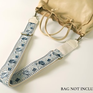 Puede incluir: Una correa de bolso desmontable con un intrincado bordado floral azul y blanco sobre una base de tela clara. La correa incluye herrajes metálicos ajustables y extremos de piel sintética blanca con clips, junto a un bolso beige.