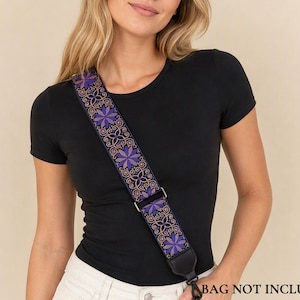 Könnte beinhalten: Ein Umhängetaschenriemen mit dunkelblauer Basis und einem sich wiederholenden Muster aus violetten sternförmigen Blumen sowie filigranen goldfarbenen Schnörkeln. Der Riemen verfügt über Metallbeschläge und schwarze Endstücke aus Kunstleder.