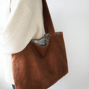 May include: A brown corduroy tote bag with a wide shoulder strap. The bag's interior lining features a floral pattern in shades of blue, yellow, and green. The bag is a large size, suitable for carrying various items.