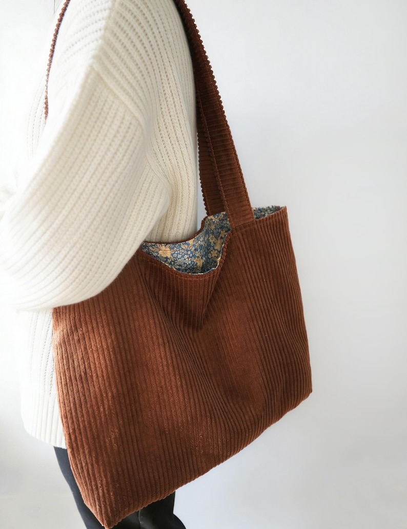 May include: A brown corduroy tote bag with a wide shoulder strap. The bag's interior lining features a floral pattern in shades of blue, yellow, and green. The bag is a large size, suitable for carrying various items.