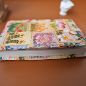 Puede incluir: Un libro de tapa dura con una cubierta estampada con flores. La cubierta presenta un dise&ntilde;o de patchwork con cuadrados de flores rosas, amarillas y naranjas. El libro est&aacute; cerrado y apoyado sobre una superficie marr&oacute;n.