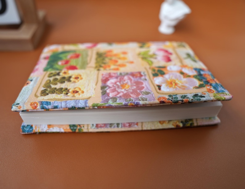 Puede incluir: Un libro de tapa dura con una cubierta estampada con flores. La cubierta presenta un dise&ntilde;o de patchwork con cuadrados de flores rosas, amarillas y naranjas. El libro est&aacute; cerrado y apoyado sobre una superficie marr&oacute;n.