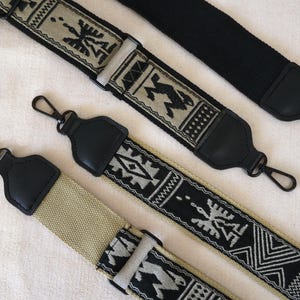 Könnte beinhalten: Drei verstellbare Taschengurte mit schwarzen lederartigen Endstücken und drehbaren Metallhaken. Die Gurte zeigen gewebte Muster in Schwarz, Beige und Weiß, darunter geometrische Formen und stilisierte vogelartige Figuren auf Stoff.