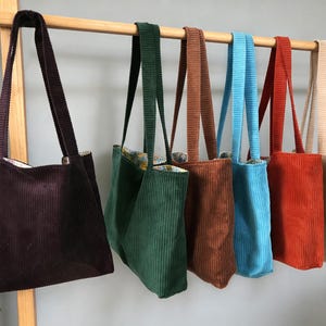 May include: A collection of corduroy tote bags in various colors, including deep purple, green, brown, blue, red-orange, and beige. Each bag has a long strap and a rectangular shape, hanging from a wooden rod.